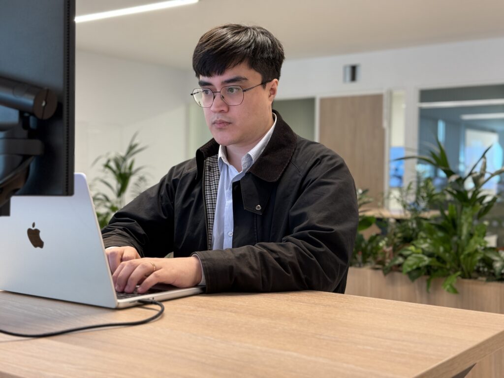 A man wearing glasses working on a laptop at a desk in a modern office setting.